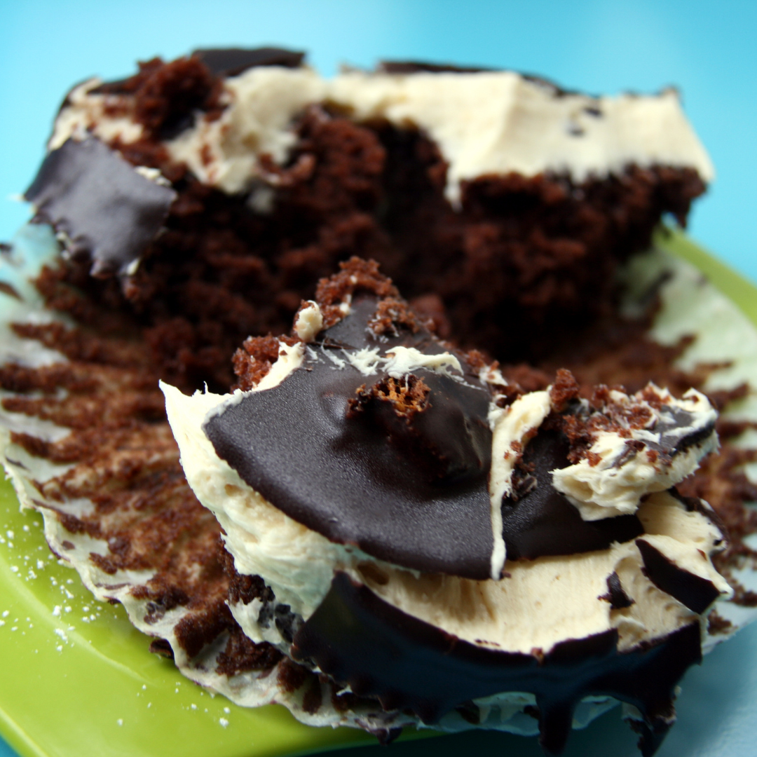 A delightfully smashed chocolate cupcake with vanilla frosting and a hard chocolate top sitting on a lime plate on a turquois background. Delicious is how it looks.